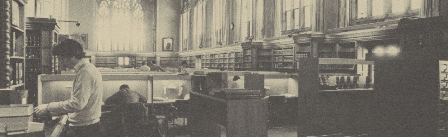 Student reading at a standing desk in the library reading room, surrounded by carrels. 