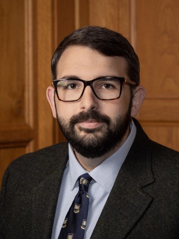 A bespectacled man looking at the camera in front of wood paneling.