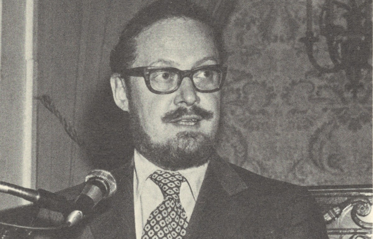 Man in a suit and glasses with a short beard speaking at a podium