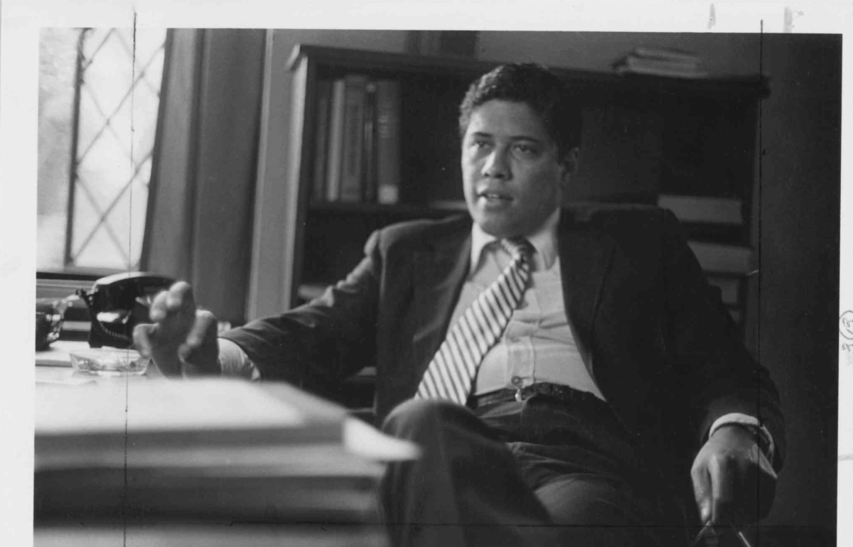 Black and white image of a man in a suit reclining with his arched hand on a desk.