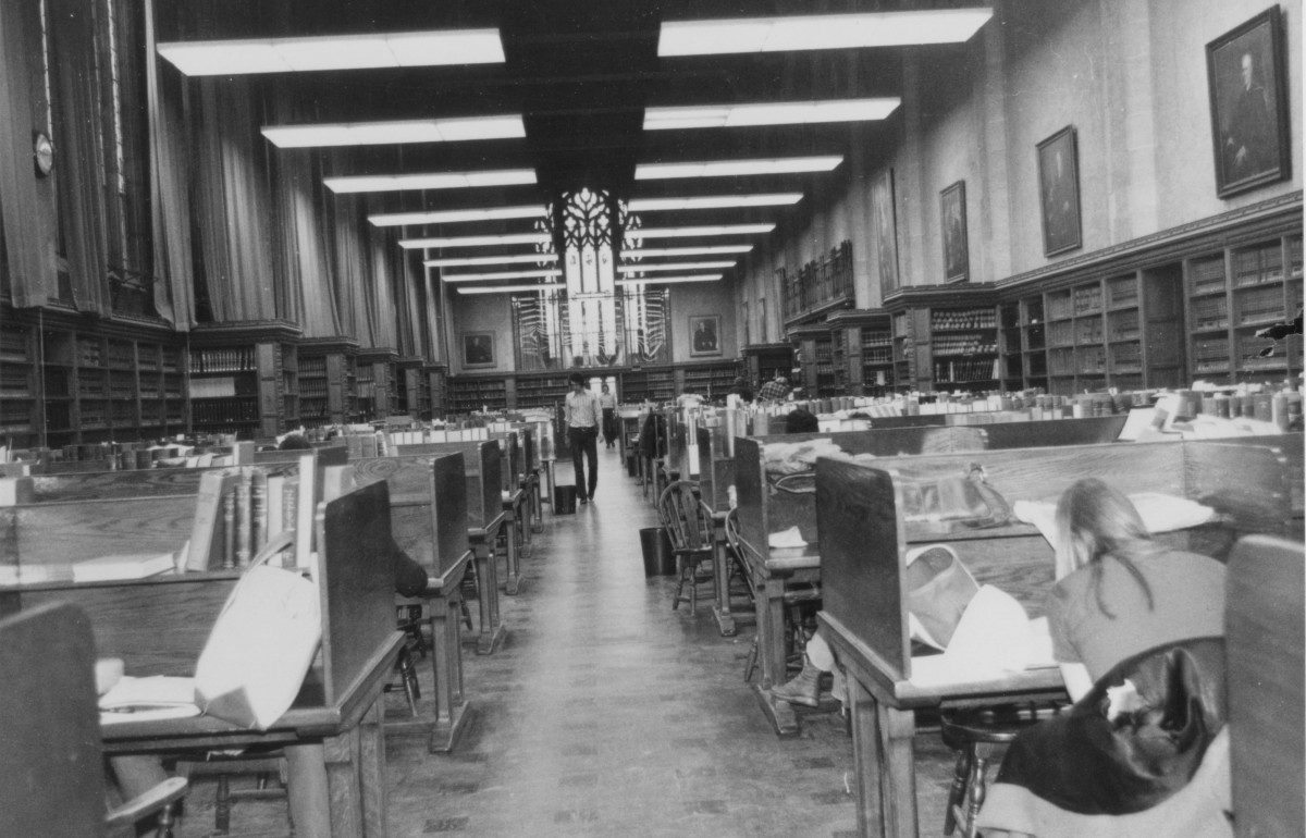 Black and white photograph of desks and student in a library.