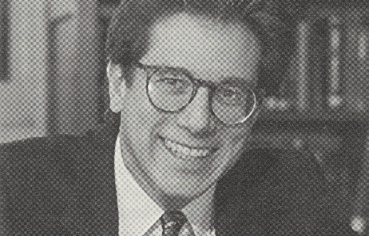 Black and white photograph of a suited man in glasses at a desk.