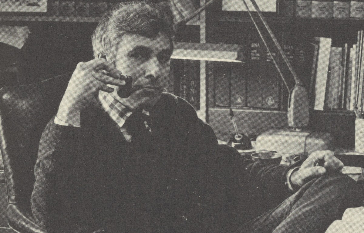 Black and white photograph of a man in a sweater smoking a pipe casually in a chair.