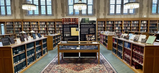 Wideshot photograph of an exhibit case with two posters behind it. Surrounded by bookcases on all sides.